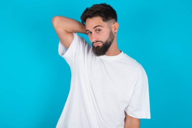 Beyaz tişört giyen beyaz sakallı beyaz bir adam mavi arka planda sırt ve boyun ağrısından muzdarip, boynuna elle dokunan, kas ağrısı olan biri..