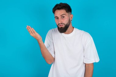 Studio shot of frustrated bearded caucasian man wearing white T-shirt over blue background gesturing with raised palm, frowning, being displeased and confused with dumb question.