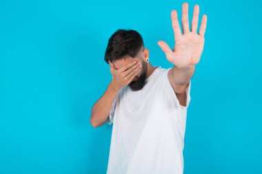Mavi arka planda beyaz tişört giyen beyaz sakallı beyaz adam avuçla gözlerini kapatıyor ve dur hareketi yapıyor, saklanmaya çalışıyor. Bana bakma, görmek istemiyorum, utanıyorum ya da korkuyorum..