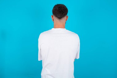 Beyaz tişörtlü beyaz sakallı beyaz bir adam mavi arka planda dikilip kollarını vücuduna dikiyor..