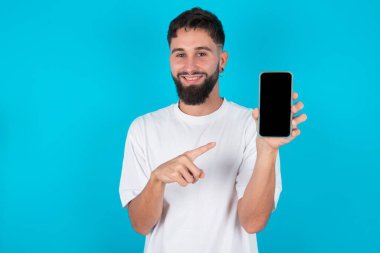 Mavi arka planda sıradan tişörtler giyen, gülümseyen beyaz adam boş telefon ekranını işaret ediyor. Reklam ve iletişim konsepti.