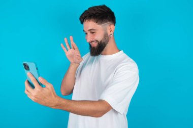 Mavi arka plan üzerine günlük tişörtler giyen mutlu ve arkadaş canlısı beyaz adamın portresi. Selfie çekip el sallıyor, video görüşmesiyle iletişim kuruyor, internette sohbet ediyor..