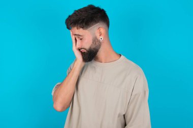Mavi arka plan üzerine günlük tişört giyen sakallı beyaz bir adam. Ağlarken yüzünü ellerle kapatan üzgün bir ifade. Depresyon kavramı.