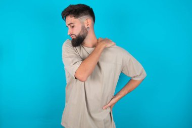 Mavi arka planda günlük tişört giyen beyaz sakallı bir adam sırt ağrısı çekiyor.