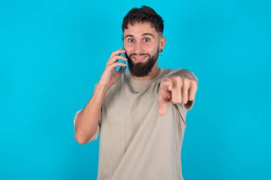 Mavi arka plan üzerine gündelik tişört giyen olumlu sakallı beyaz adam doğrudan kameraya bakıyor telefon konuşmaları uzun saatler konuşmaktan keyif alıyor. Bana katıl.