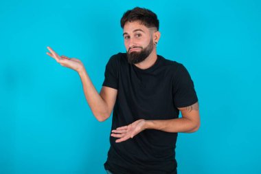 Siyah tişörtlü genç, İspanyol sakallı adam mavi arka planda duruyor iki elinde garip bir şey var ve 