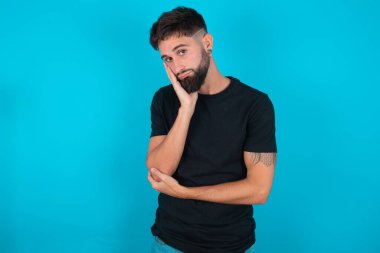 Çok sıkılmış İspanyol sakallı bir adam, siyah tişört giyip mavi arka planda el ele tutuşurken diğer eli çapraz tutarak yorgun ve hasta görünüyor.,