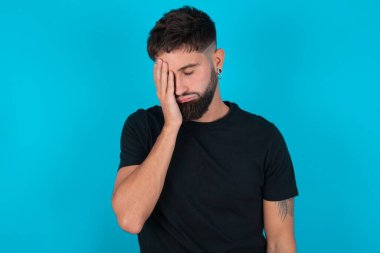 Mavi arka planda duran siyah tişört giyen yorgun ve fazla çalışan İspanyol sakallı genç adamın uykulu bir ifadesi, kasvetli bir bakışı var, yüzü elle örtülmüş, gözleri kapalı, yorgunluktan nefesi kesilmiş, partiden sonra bitkinlik ve yorgunluk var.