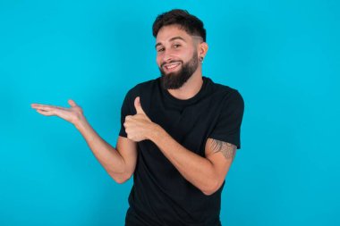 Siyah tişört giyen genç İspanyol sakallı adam mavi arka planda duruyor, avuç içini gösteriyor ve baş parmaklarıyla hareket ediyor, mutlu ve neşeli gülümsüyor..