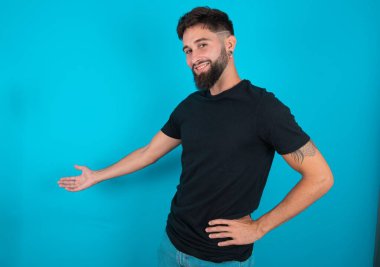 Siyah tişört giyen genç İspanyol sakallı adam mavi arka planda duruyor. Mutlu ve neşeli hissediyor, gülümsüyor ve seni hoş karşılıyor. Dostça bir jestle içeri davet ediyor.