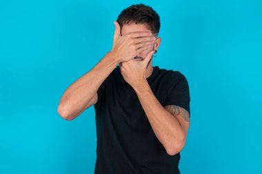 Siyah tişört giyen genç İspanyol sakallı adam mavi arka planda duruyor gözleri ve ağzı kapalı, şaşırmış ve şok olmuş. Duyguları gizlemek.