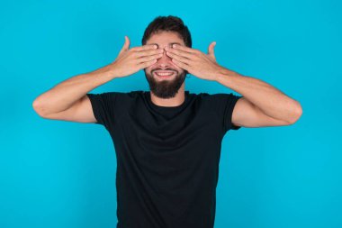 Siyah tişört giyen genç İspanyol sakallı adam mavi arka planda duruyor. Gözleri kapayarak neşeli ve komik gülümsüyor. Kör kavram.