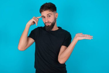 Siyah tişört giyen genç, İspanyol sakallı adam mavi arka planda duruyor. Avuç içi açık, fotokopi alanı gösteren ve alnını işaret eden avuçlar açık. Bunu bir düşün..