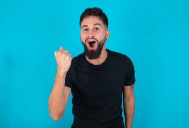 Siyah tişört giyen genç İspanyol sakallı adam mavi arka planda kızgın ve kızgın duruyor öfkeyle bağırırken yumruğunu kaldırıyor. Öfke ve agresif kavram.