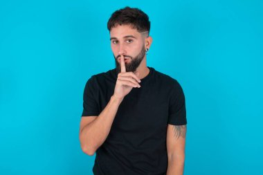 Mavi arka planda duran siyah tişörtlü İspanyol sakallı adam sessiz hareketler yapıyor, parmaklarını dudaklardan uzak tutuyor. Sessizlik ve gizli konsept.
