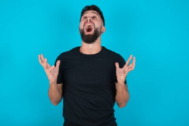 Siyah tişört giyen İspanyol sakallı adam mavi arka planda duruyor deli deli deli bağırıyor ve bağırıyor agresif bir ifade ve kollar havada. Hayal kırıklığı konsepti.