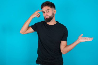 Siyah tişört giyen İspanyol sakallı adam mavi arka planda duruyor. Kafası karışmış ve avuç içi açık bir şekilde fotokopi alanı ve alnını gösteriyor. Bunu bir düşün..