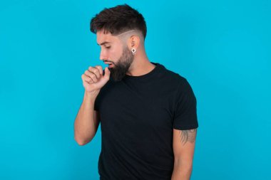 Siyah tişört giyen İspanyol sakallı bir adam mavi arka plan karşısında kendini kötü hissediyor ve soğuk algınlığı veya bronşitin belirtisi olarak öksürüyor. Sağlık hizmetleri kavramı.
