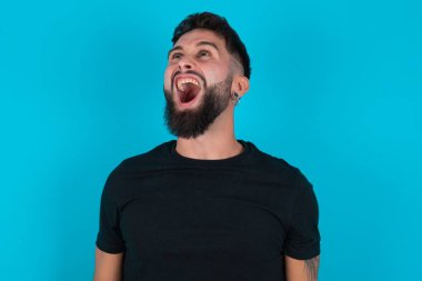 Siyah tişört giyen İspanyol sakallı adam mavi arka planda öfkeli ve öfkeli bir şekilde bağırıyor. Öfkeyle bağırıyor. Öfke ve agresif kavram.
