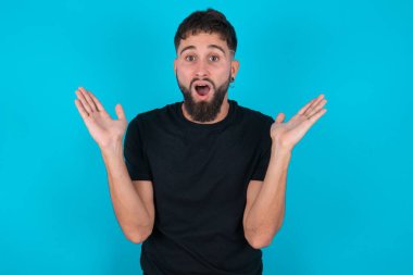 Şaşkın ve korkmuş, siyah tişörtlü İspanyol sakallı adam mavi arka plan jestleri karşısında belirsizlikle duruyor, kameraya bakıyor, şaşırmış bir şekilde hileli soruya cevap vermiyor, insanlar, vücut dili, duygu kavramı.