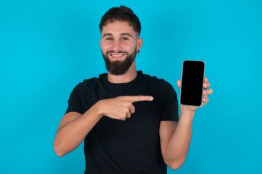 Siyah tişört giyen, gülümseyen, İspanyol sakallı adam. Mavi arka planda fotokopi alanı gibi duruyor. Boş ekran ile işaret parmağı cep telefonu üzerinde
