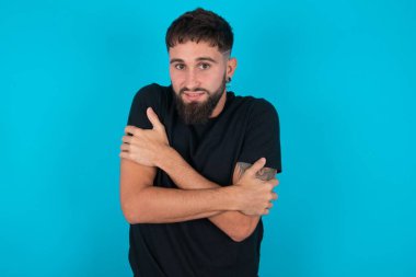 Siyah tişört giyen umutsuz İspanyol sakallı adam mavi arka planda dikiliyor ve ürperiyor, ısınmak için kendine sarılıyor ya da korkunç bir şey fark ediyor..