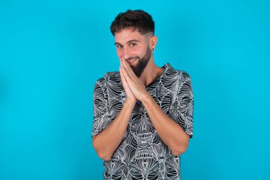 Neşeli, genç, İspanyol sakallı adam mavi arka planda durup neşeyle gülüyor ve avuçlarını birbirine bastırıp komik bir şey duyuyor.
