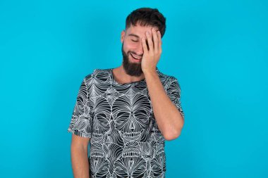 Genç İspanyol sakallı adam mavi arka planda duruyor. Avuç içi gibi gülümsüyor ve kıkırdıyor. Komik espriler duyuyor.