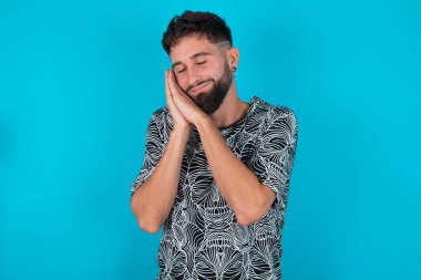 Mavi arka planda duran genç İspanyol sakallı adam avuçlarına yaslanıyor gözlerini kapıyor ve hoş bir gülümseme görüyor.