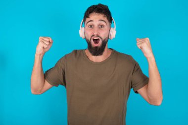 Duygulu, genç, İspanyol sakallı bir adam mavi arka planda yüksek sesle bağırıyor. Kazanan yumruklarını kaldırıyor, yumruklarını sıkıyor, ağzını açık tutuyor kulaklıklar takıyor, müzik kulaklıkları takıyor, evet jestleri yapıyor, en sevdiği müziği dinliyor.