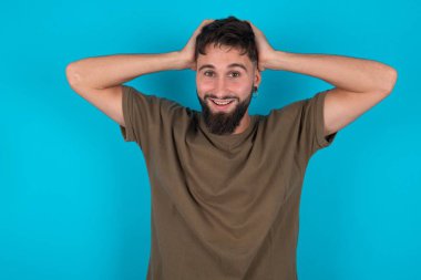 Neşeli, neşeli, genç, İspanyol sakallı adam mavi arka planda duruyor. Harika haberler aldıktan sonra ellerini havaya kaldırıyor..