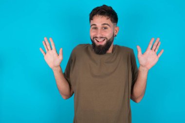 Mavi arka planda duran mutlu, genç, İspanyol sakallı adam harika bir sonuç elde ettikten sonra ağzını ve kollarını açar.