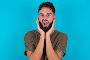 Üzgün genç, İspanyol sakallı adam mavi arka planda duruyor iki eliyle yüzüne dokunuyor.