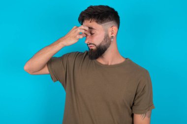 Üzgün, genç, İspanyol sakallı adam mavi arka planda duruyor. Baş ağrısından acı çekiyor.