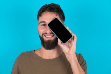 Genç İspanyol sakallı adam mavi arka planda duruyor. Elinde modern akıllı telefon var. Bir gözünü kapatıp gülümsüyor.