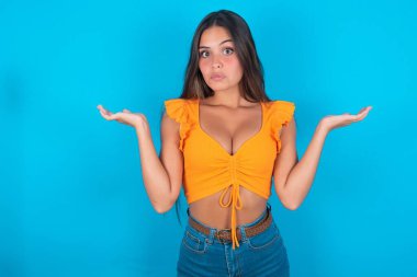 Kafası karışmış ve cahil, esmer, turuncu bluz giyen, kolları açık, omuzlarını silkmiş, 