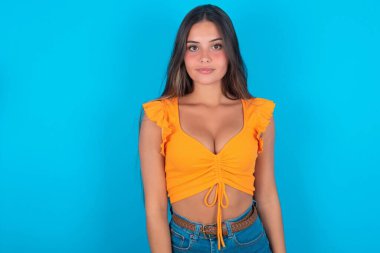 Mavi arka planda turuncu kolsuz bluz giyen neşeli, güzel esmer kadın kameraya bakıyor, bir şey düşünüyor. İki kolunu da indir, tarafsız yüz ifadesi..