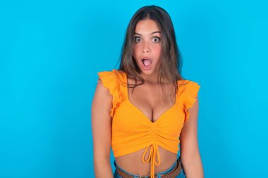 Etkileyici yüz ifadeleri. Şok olmuş güzel esmer kadın mavi arka planda turuncu kolsuz bluz giyiyor. Gördüklerinden sersemlemiş hissediyor..