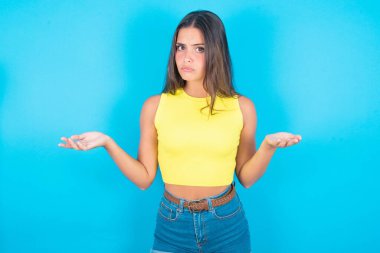 Mavi arka planda sarı kolsuz bluz giyen güzel esmer kadın belirsiz omuz silkiyor..