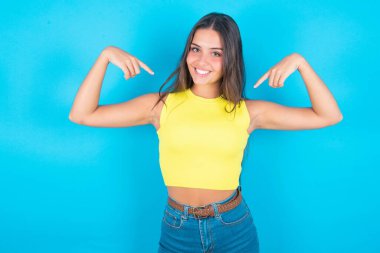 Beni seç! Kendine güvenen, kendine güvenen, karizmatik, güzel, sarı kolsuz bluz giyen, mavi arka planda kendini terfi ettiren, geniş bir gülümseme isteyen ve vücuduna işaret eden bir kadın..