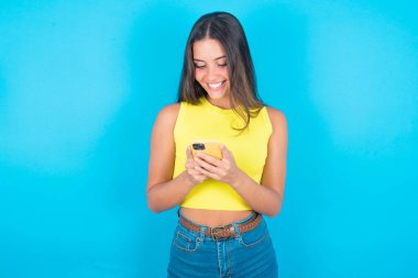 Mavi arka planda sarı kolsuz bluz giyen güzel esmer kadın cep telefonu kullanıyor, mesajlaşıyor, arkadaşlarıyla mesajlaşıyor, akıllı telefona bakıyor. Modern teknolojiler ve iletişim.