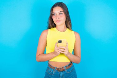 Mavi arka planda sarı atlet giyen güzel, mutlu bir esmer kadın cep telefonu kullanarak kulaklıkla müzik dinliyor..