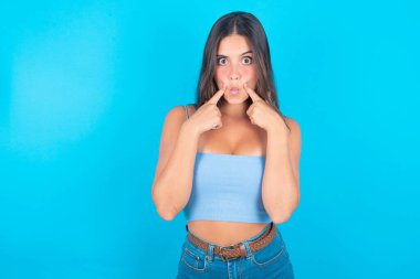 Mavi kolsuz bluz giyen neşeli, genç esmer kadın mavi arka planda balık dudakları yapıyor.  