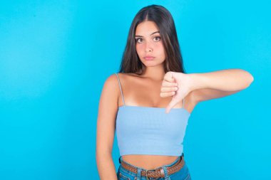 Mavi arka planda mavi kolsuz bluz giyen genç esmer kadın mutsuz ve kızgın görünüyor. Reddedilmiş ve olumsuz..