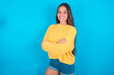 Sarı süveter giyen esmer bir kadının portresi mavi arka planda duruyor, kolları bağlı ve gülümsüyor.