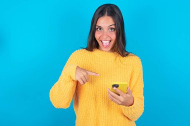 Mavi arka planda sarı süveter giyen akıllı telefonlu sevimli, şaşkın, genç esmer kadın.