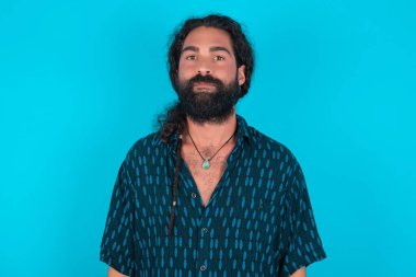 Mavi arka planda mavi gömlekli neşeli beyaz adam kameraya bakıyor, bir şey düşünüyor. İki kolunu da indir, tarafsız yüz ifadesi..