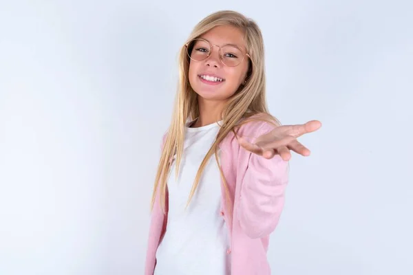 Blondynka Różowej Kurtce Okularach Białym Tle Rozciągająca Dłoń Aparatu Uśmiechnięta — Zdjęcie stockowe
