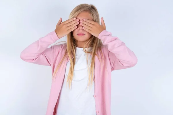 手で目を覆う白い背景の上にピンクのジャケットとメガネを身に着けているブロンドの少女 — ストック写真
