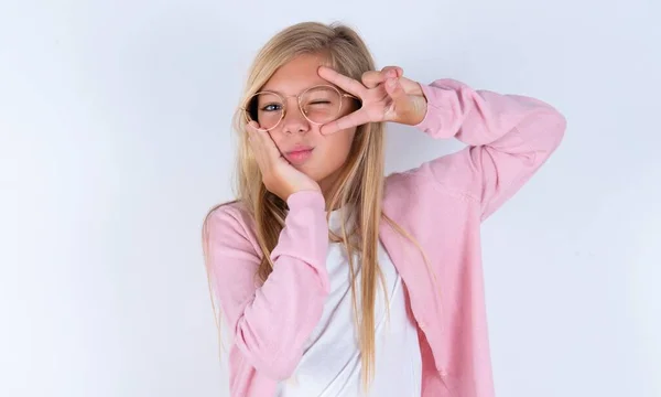 金发小女孩 身穿粉色夹克 戴着眼镜 罩在白色背景上 近视时做V字形标志 — 图库照片
